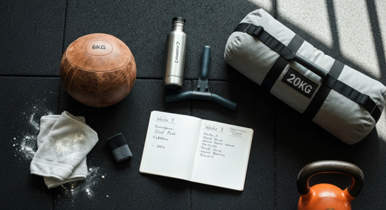 Flatlay mit Hyrox-Equipment - Wall Ball, Sandbag, Ruder-Griff, Trainings-Notizbuch